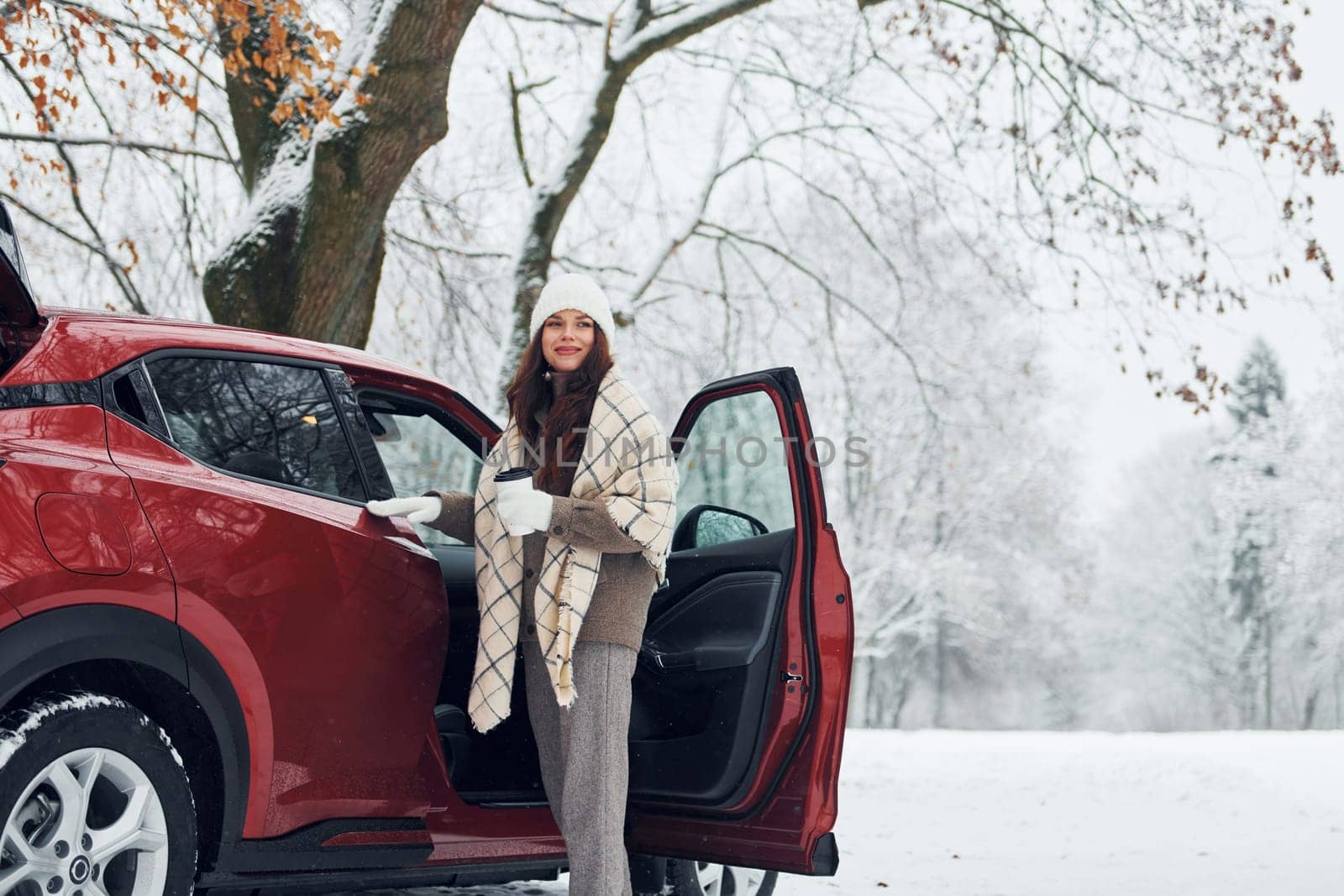 Beautiful young woman is outdoors near her red automobile at winter time.