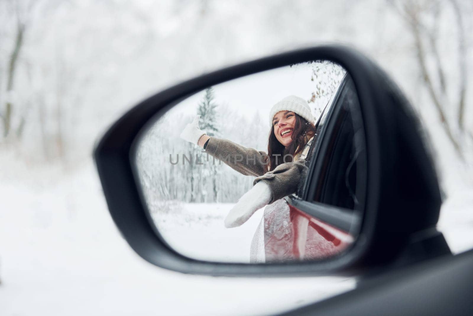 View in the side mirror. Beautiful young woman is outdoors near her red automobile at winter time.
