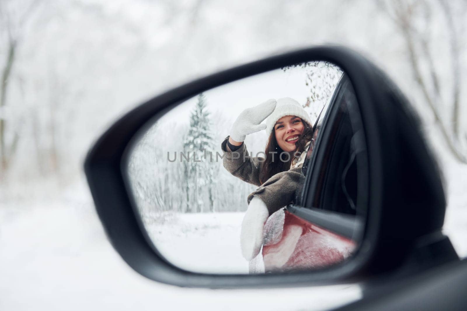 View in the side mirror. Beautiful young woman is outdoors near her red automobile at winter time.