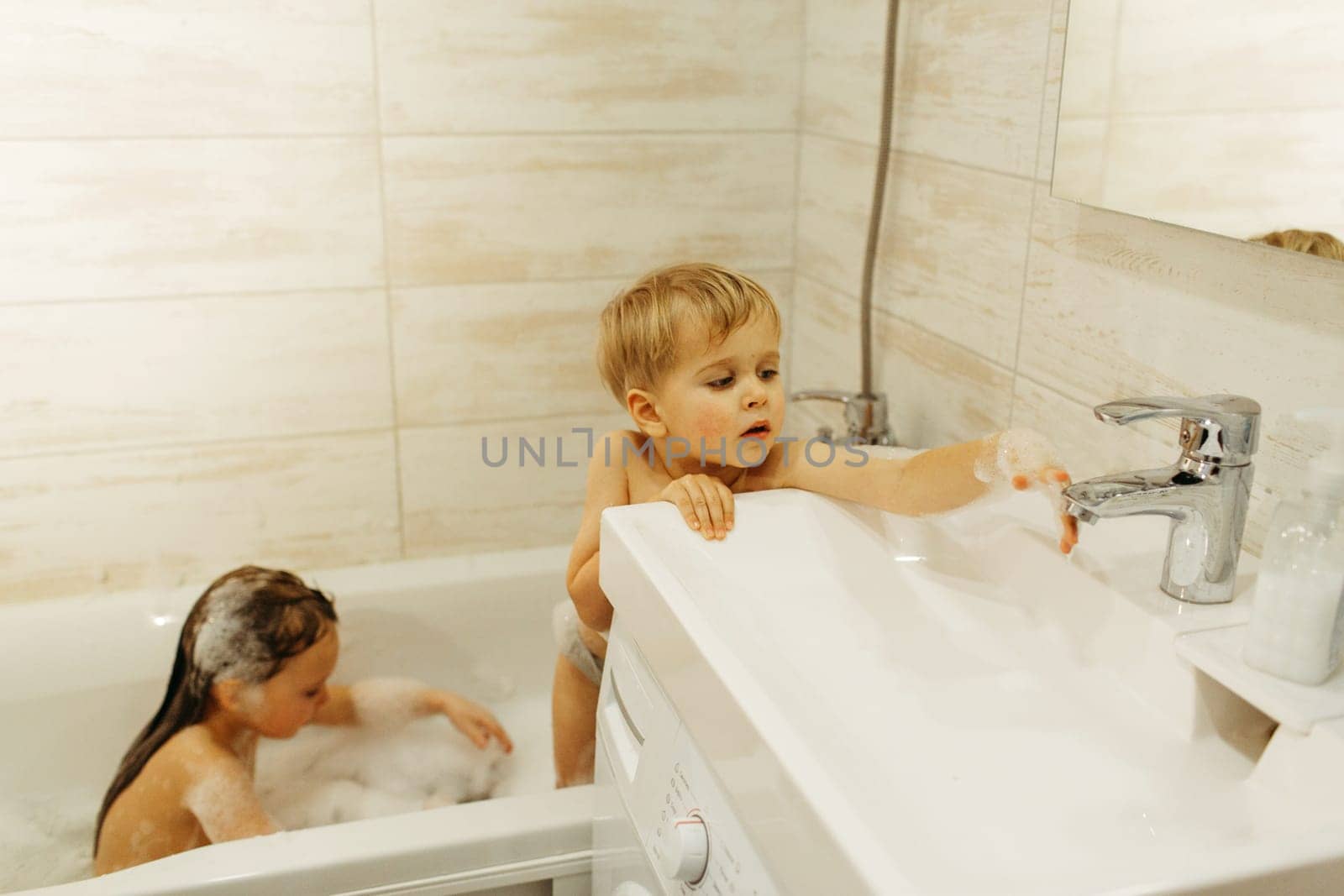 A girl and a boy of preschool age bathe in the bathroom. A girl plays with foam, a boy with water flowing from a tap.