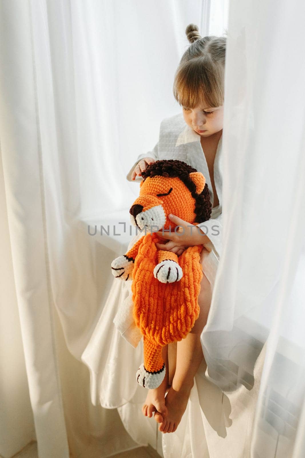 A girl in a bathrobe plays on the windowsill with a knitted lion.