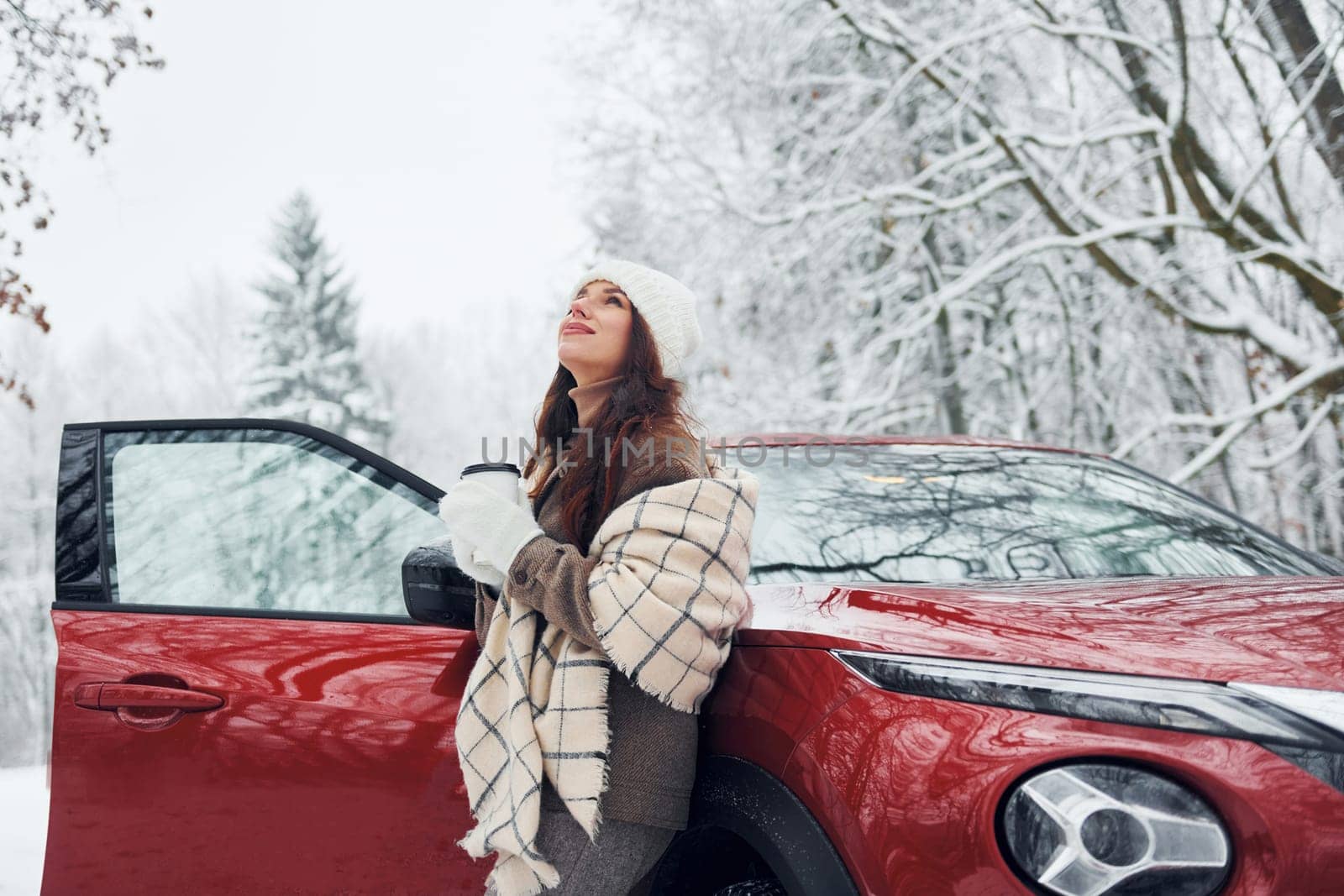 Non urban scene. Beautiful young woman is outdoors near her red automobile at winter time.