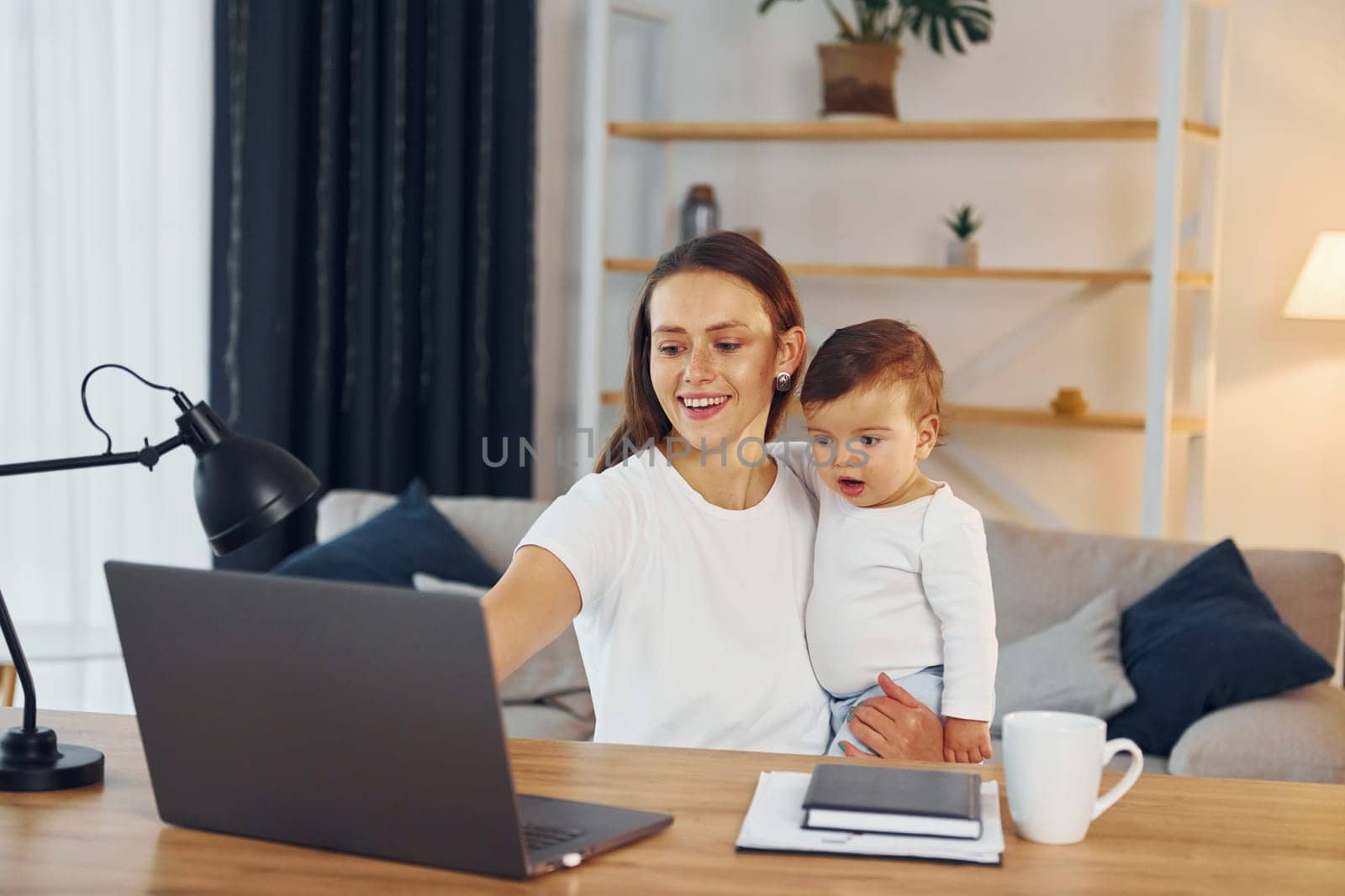 Sitting by the table with laptop. Mother with her little daughter is at home together.