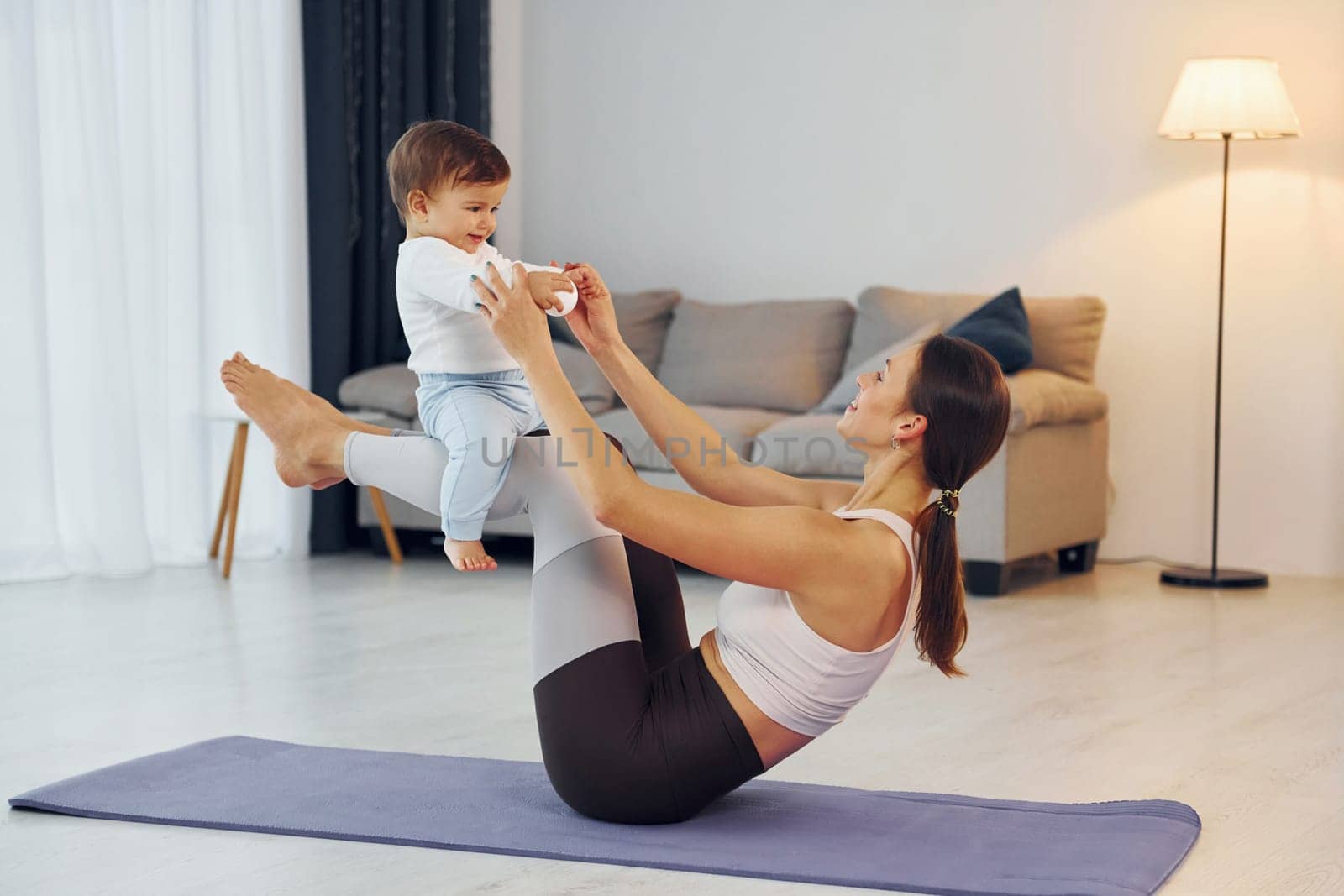 Woman laying down on mat and holding little girl. Mother with her little daughter is at home together by Standret