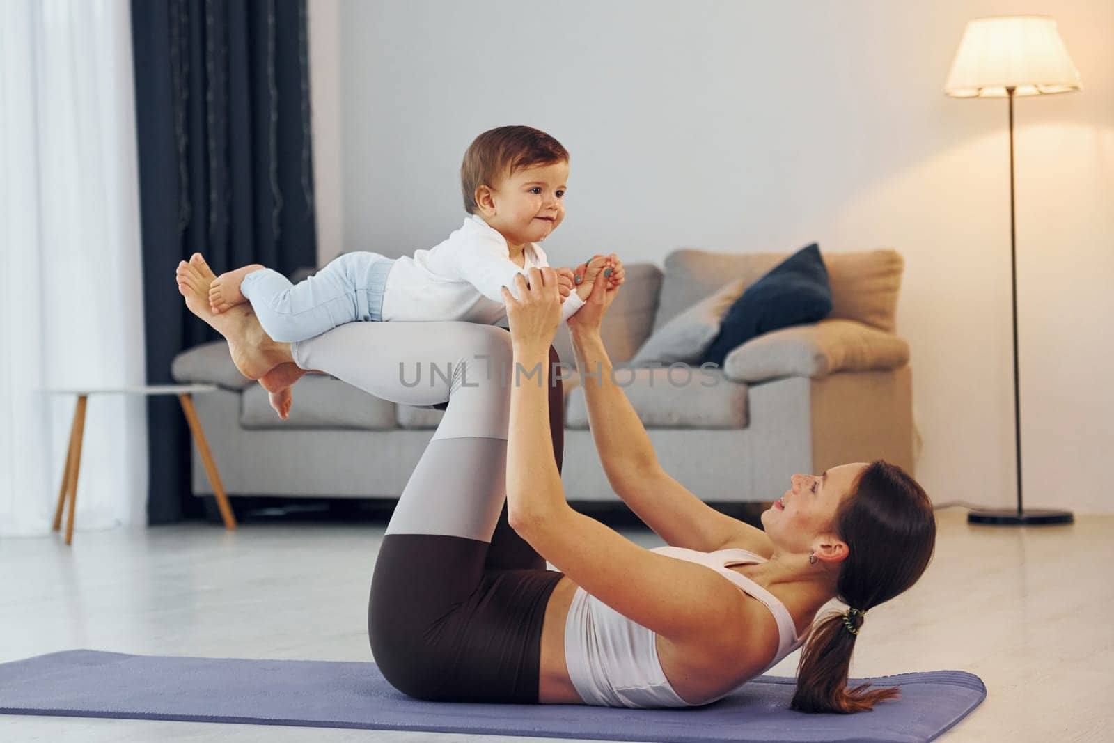 Woman laying down on mat and holding little girl. Mother with her little daughter is at home together by Standret