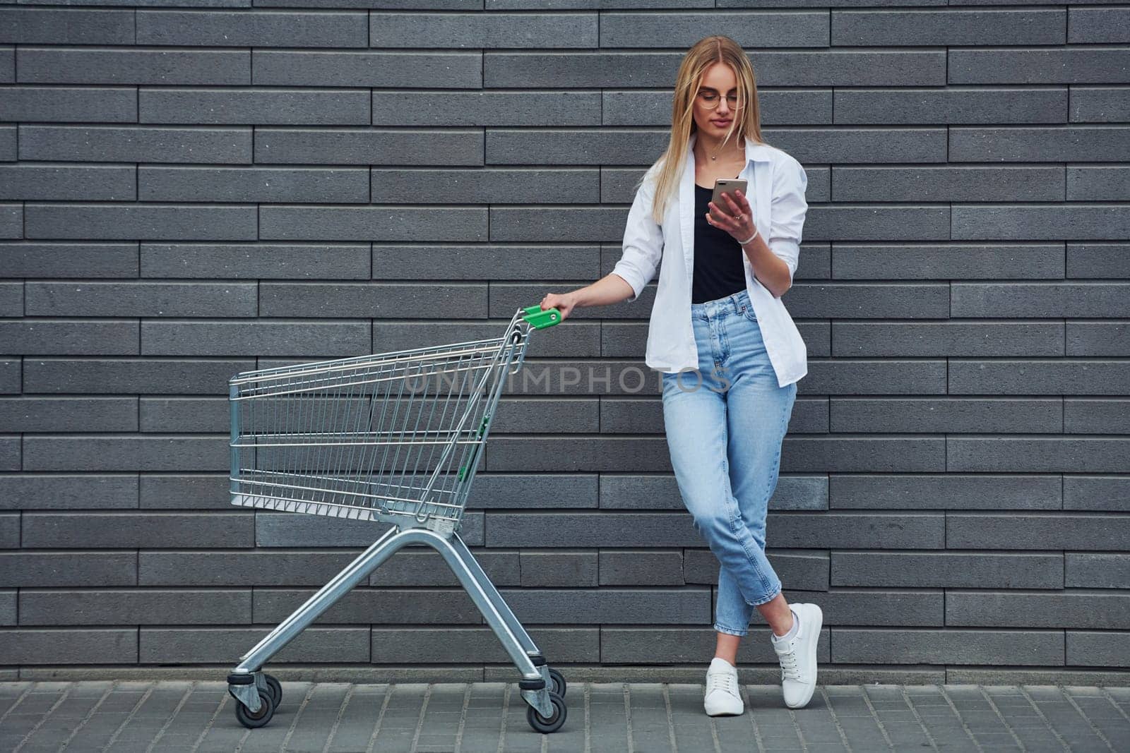 Beautiful blonde in casual clothes with shopping cart is outdoors at sunny daytime.