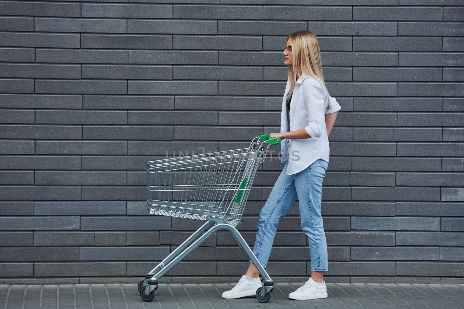 Beautiful blonde in casual clothes with shopping cart is outdoors at sunny daytime by Standret