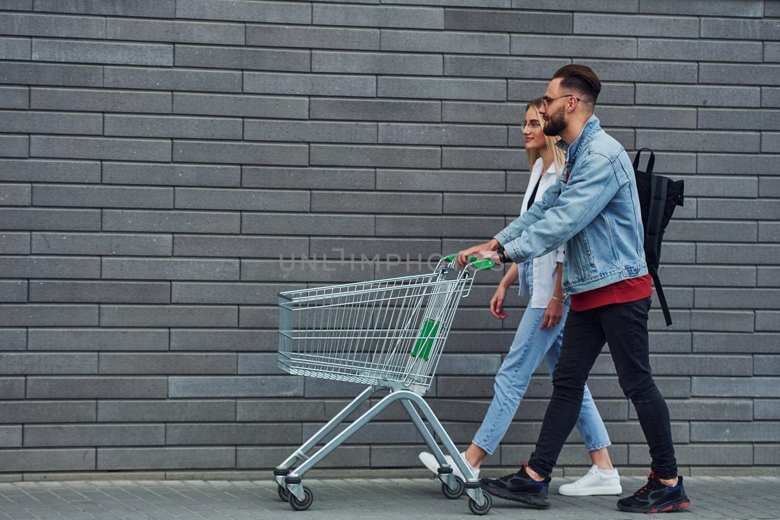 With shopping cart. Young stylish man with woman in casual clothes outdoors together. Conception of friendship or relationships.