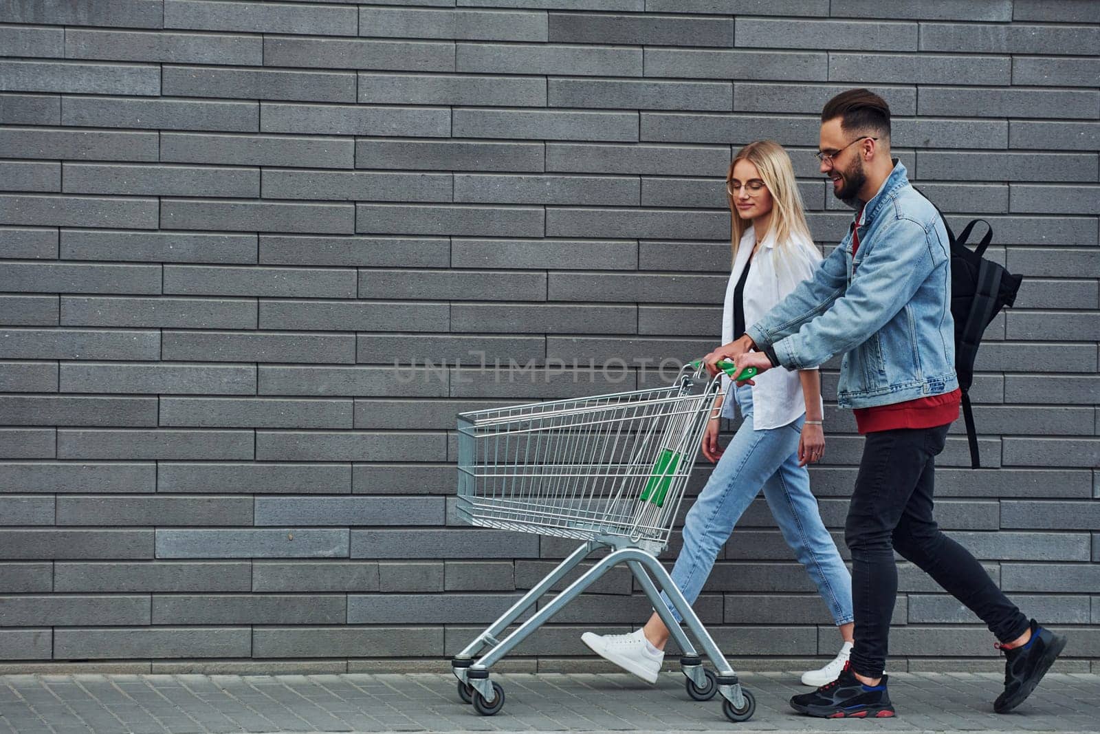 With shopping cart. Young stylish man with woman in casual clothes outdoors together. Conception of friendship or relationships.