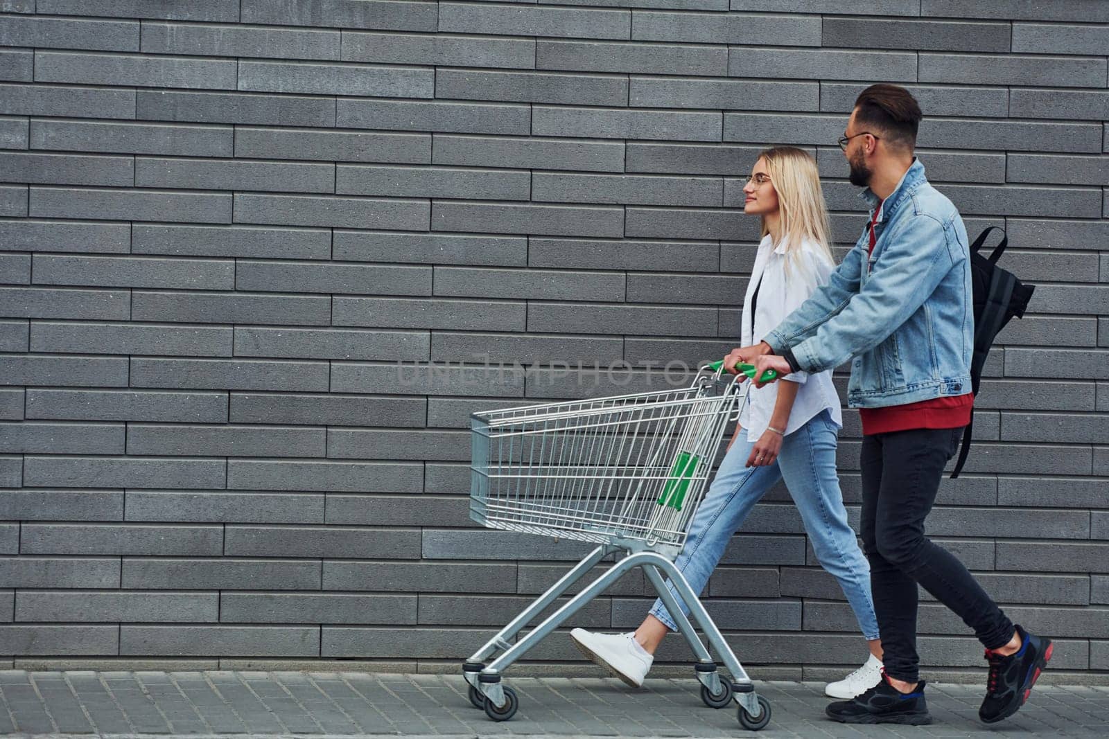 With shopping cart. Young stylish man with woman in casual clothes outdoors together. Conception of friendship or relationships.