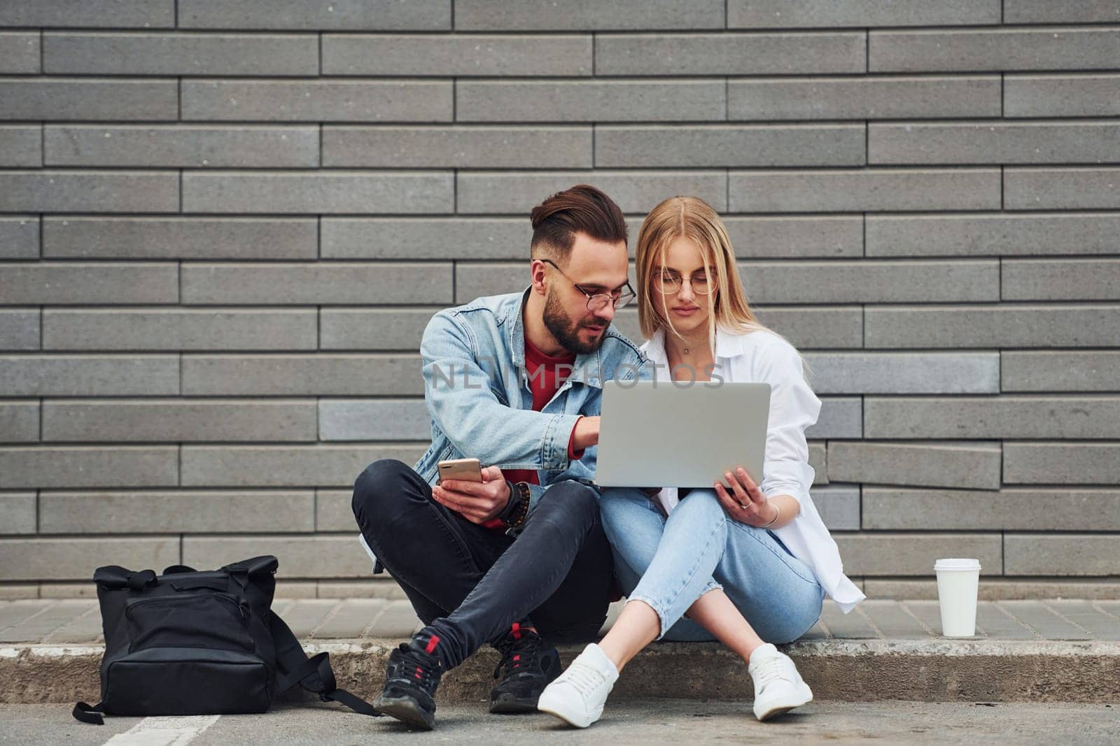 Using laptop. Young stylish man with woman in casual clothes sitting outdoors together. Conception of friendship or relationships.