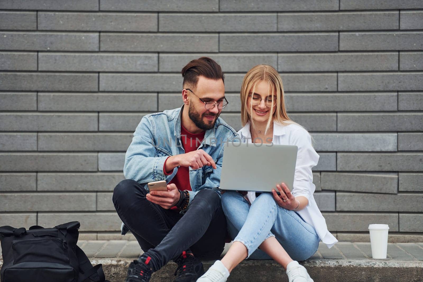 Using laptop. Young stylish man with woman in casual clothes sitting outdoors together. Conception of friendship or relationships.