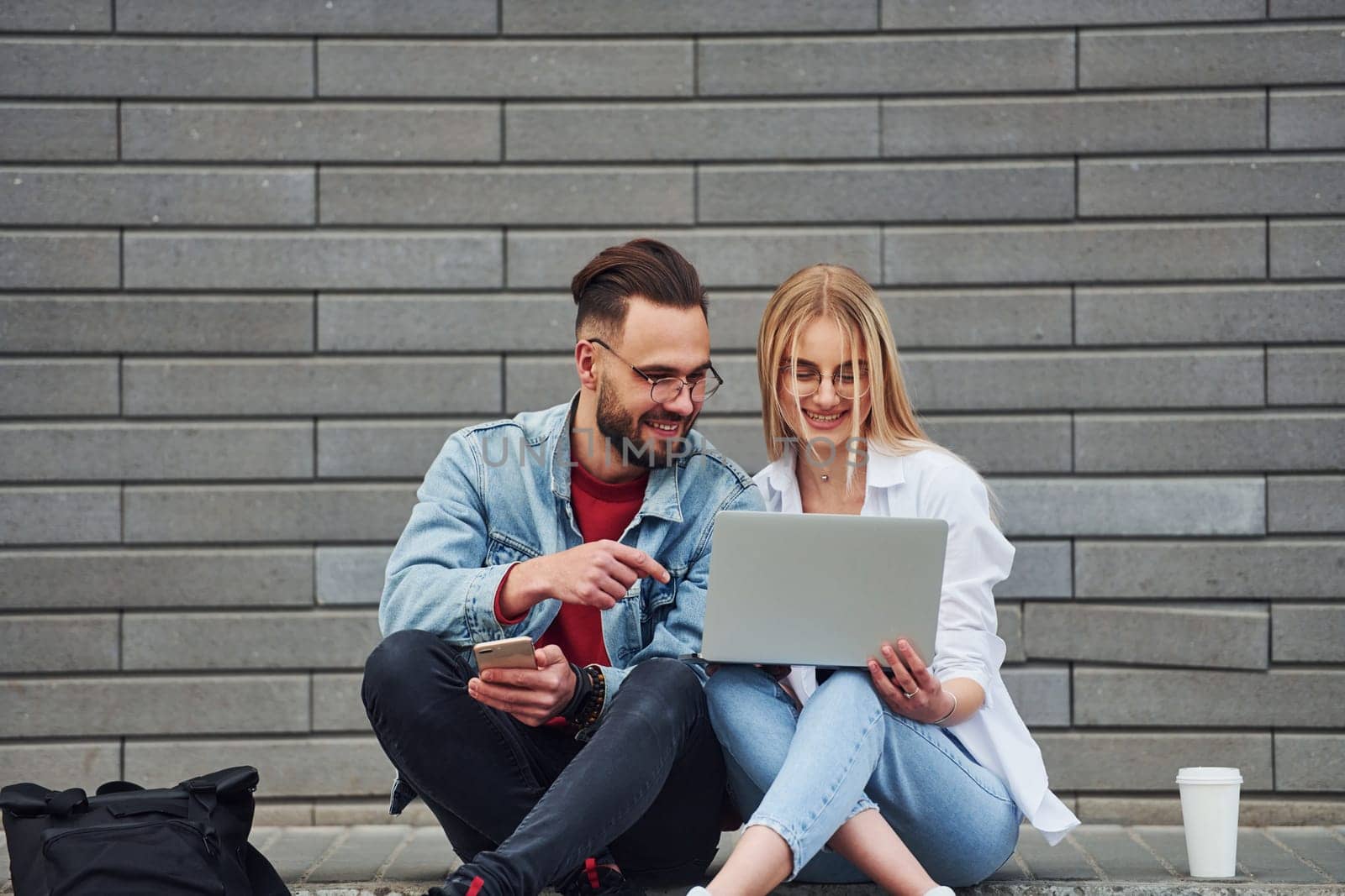 Using laptop. Young stylish man with woman in casual clothes sitting outdoors together. Conception of friendship or relationships.