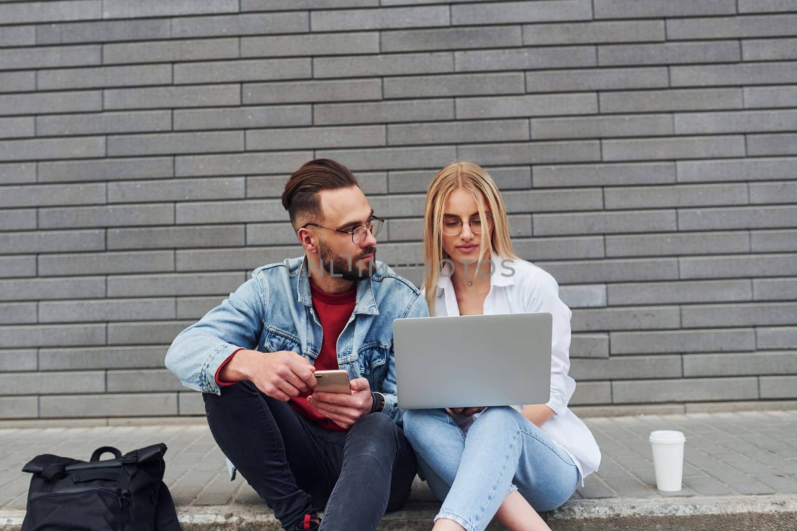 Using laptop. Young stylish man with woman in casual clothes sitting outdoors together. Conception of friendship or relationships.