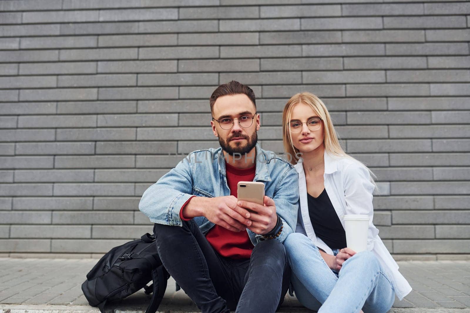 Using phone. Young stylish man with woman in casual clothes sitting outdoors together. Conception of friendship or relationships.