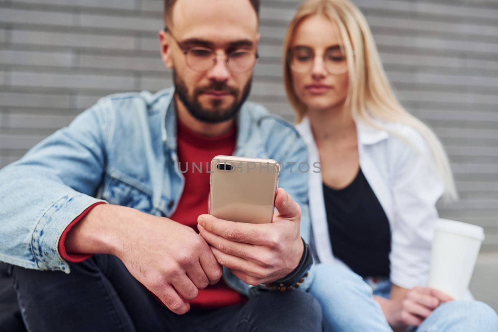 Using phone. Young stylish man with woman in casual clothes sitting outdoors together. Conception of friendship or relationships.