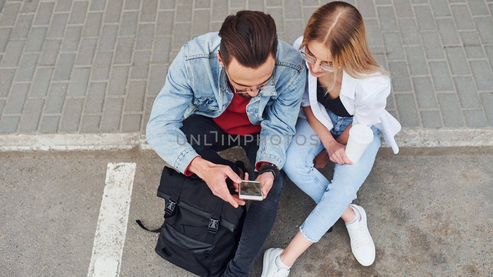 Using phone. Young stylish man with woman in casual clothes sitting outdoors together. Conception of friendship or relationships.