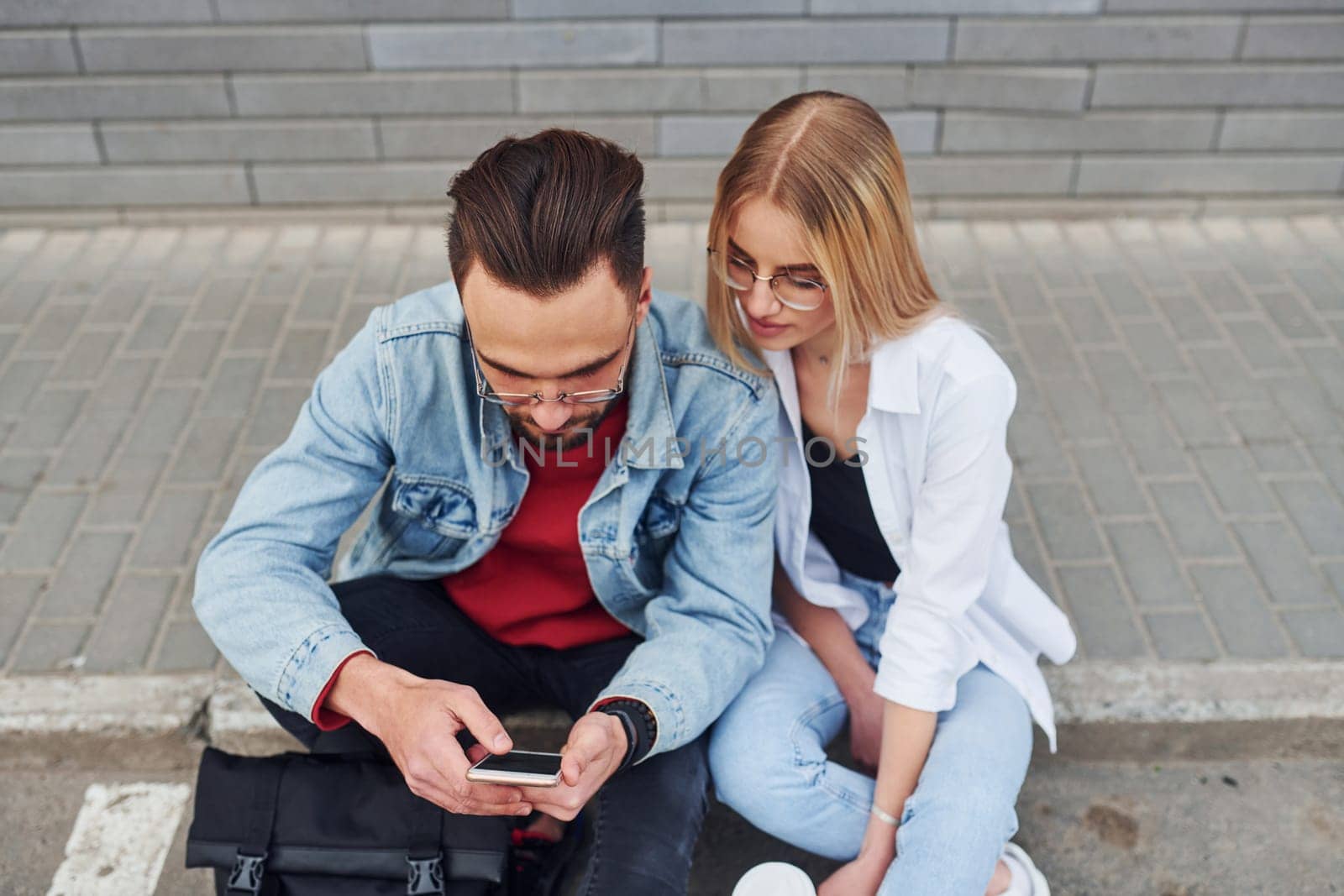 Using phone. Young stylish man with woman in casual clothes sitting outdoors together. Conception of friendship or relationships by Standret