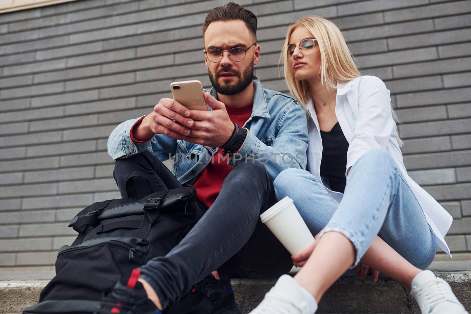 Using phone. Young stylish man with woman in casual clothes sitting outdoors together. Conception of friendship or relationships by Standret