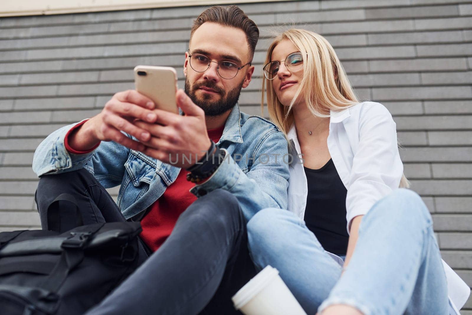 Using phone. Young stylish man with woman in casual clothes sitting outdoors together. Conception of friendship or relationships by Standret