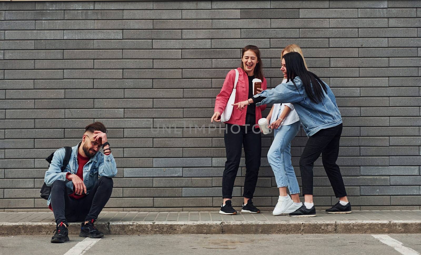 Three women standing and laughing at guy that sitting outdoors near building at daytime.