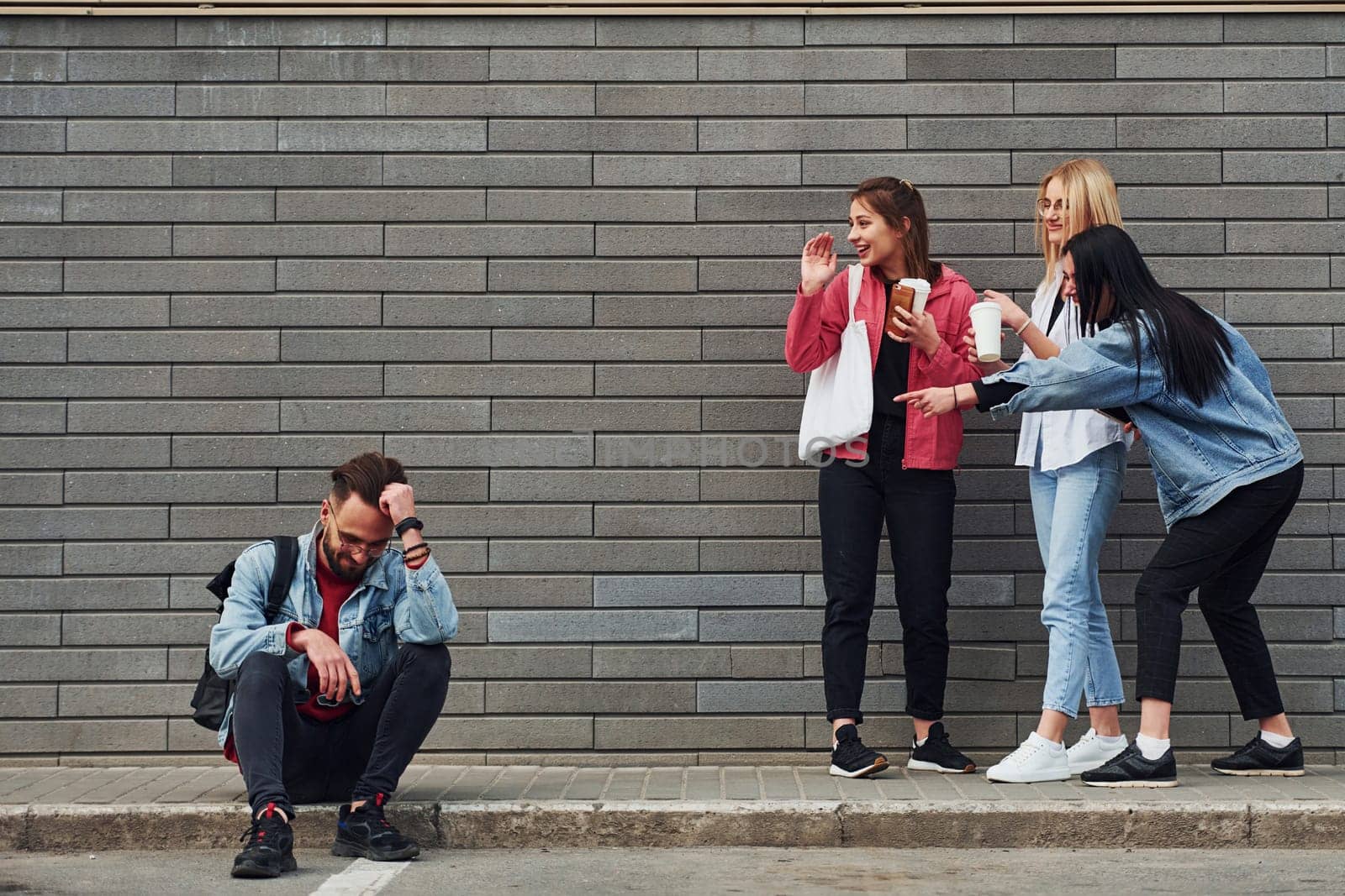 Three women standing and laughing at guy that sitting outdoors near building at daytime.