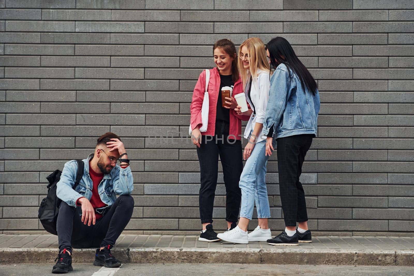 Three women standing and laughing at guy that sitting outdoors near building at daytime by Standret