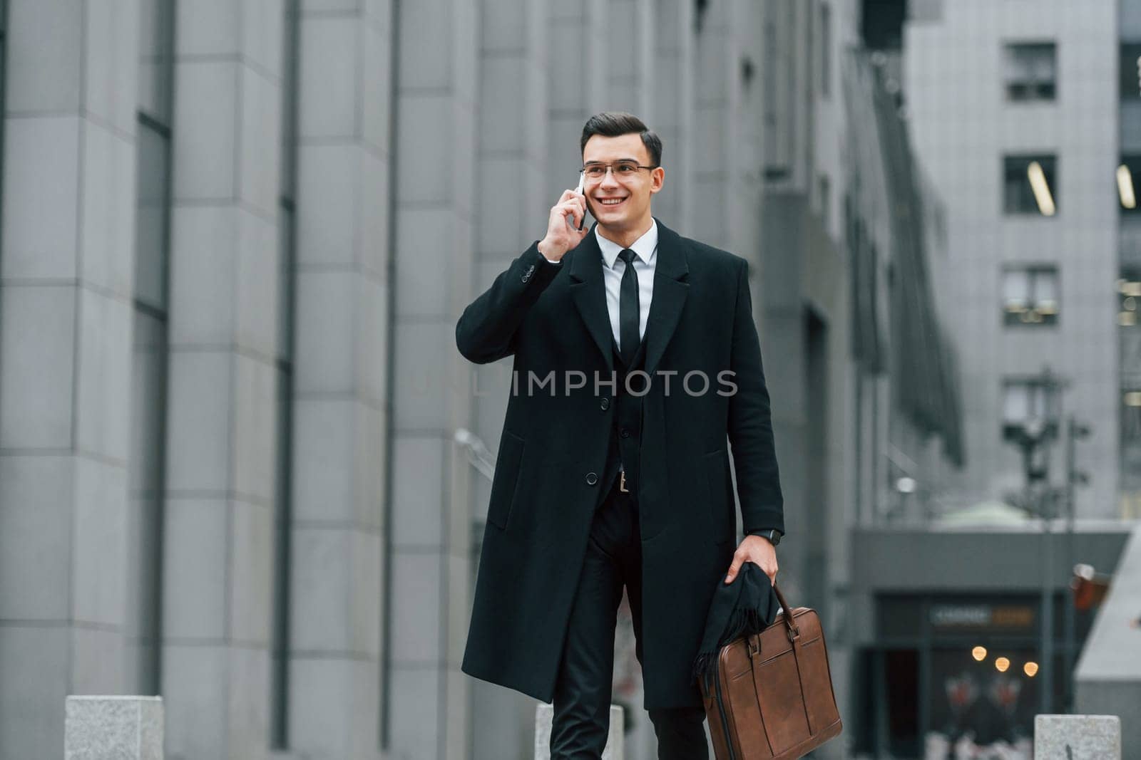 With smartphone. Businessman in black suit and tie is outdoors in the city.