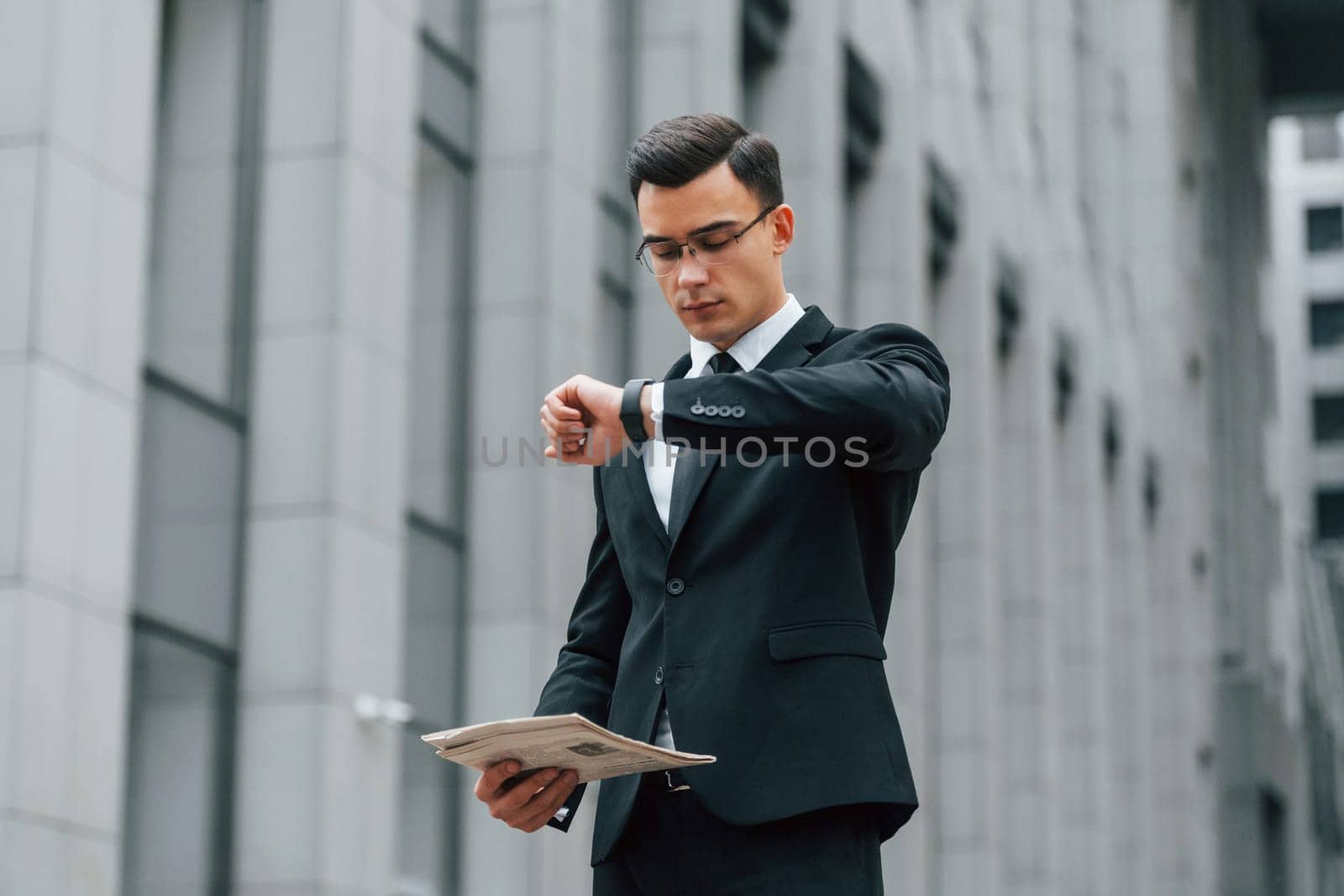 Holding newspaper. Businessman in black suit and tie is outdoors in the city.