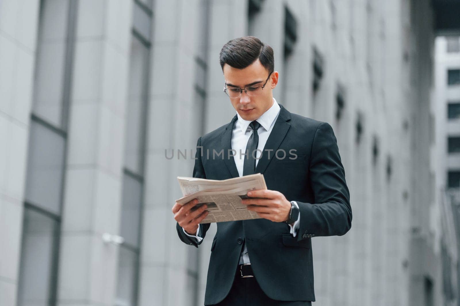 Holding newspaper. Businessman in black suit and tie is outdoors in the city.