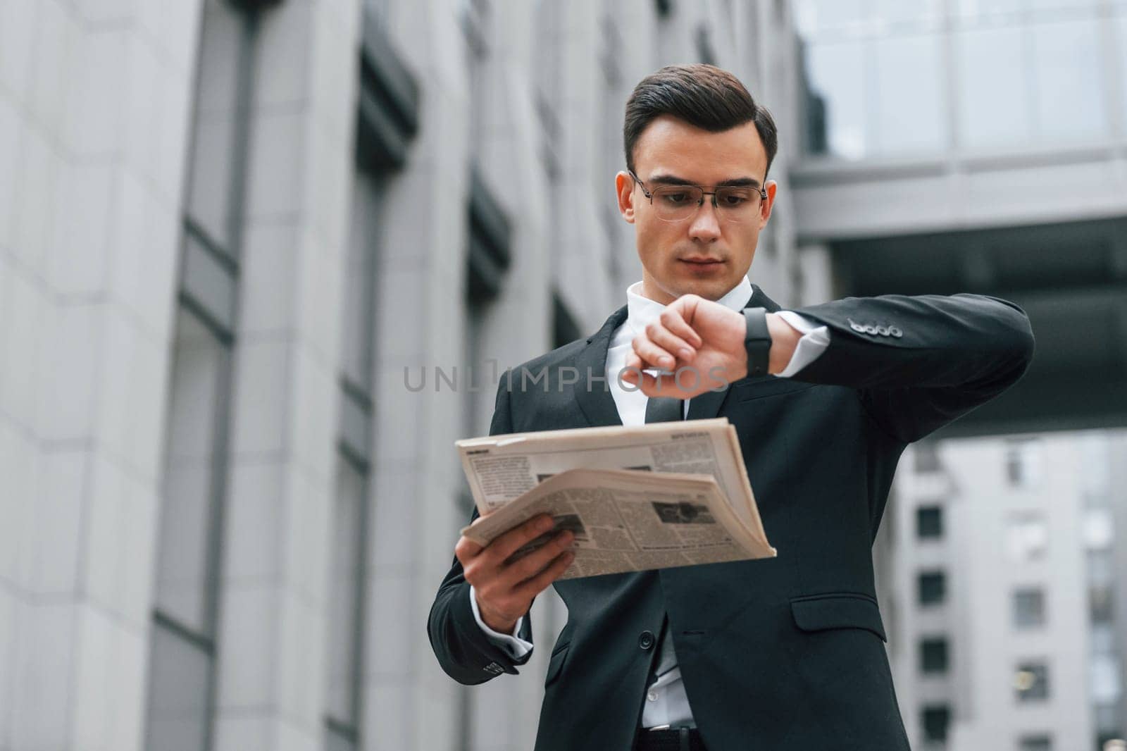Holding newspaper. Businessman in black suit and tie is outdoors in the city.