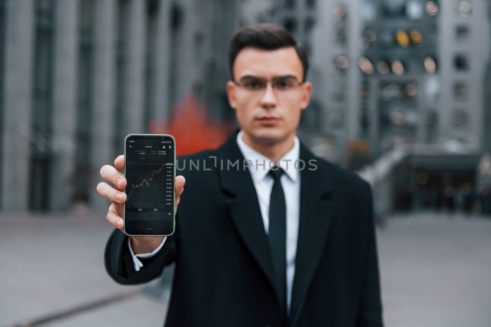 Showing phone with crypto graphs in it. Businessman in black suit and tie is outdoors in the city.