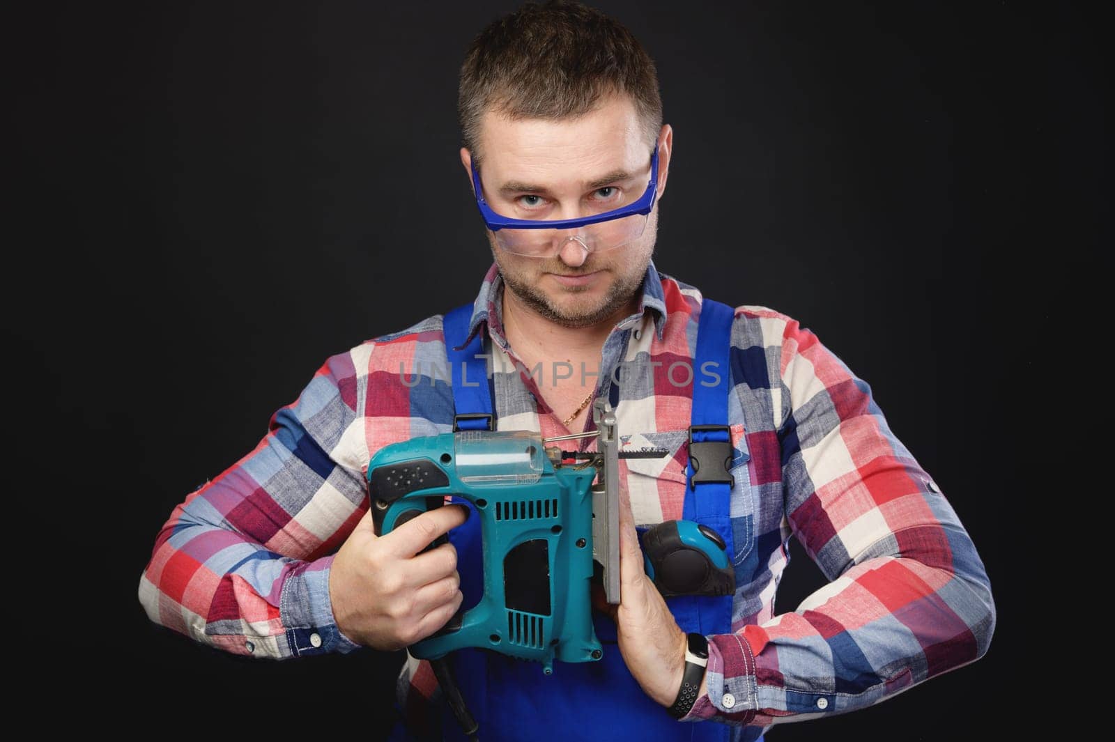 A male engineer is looking at the camera while holding a tool. Satisfied master carpenter in goggles holds a large professional construction tool in front of him. White builder posing with tools in his hands.