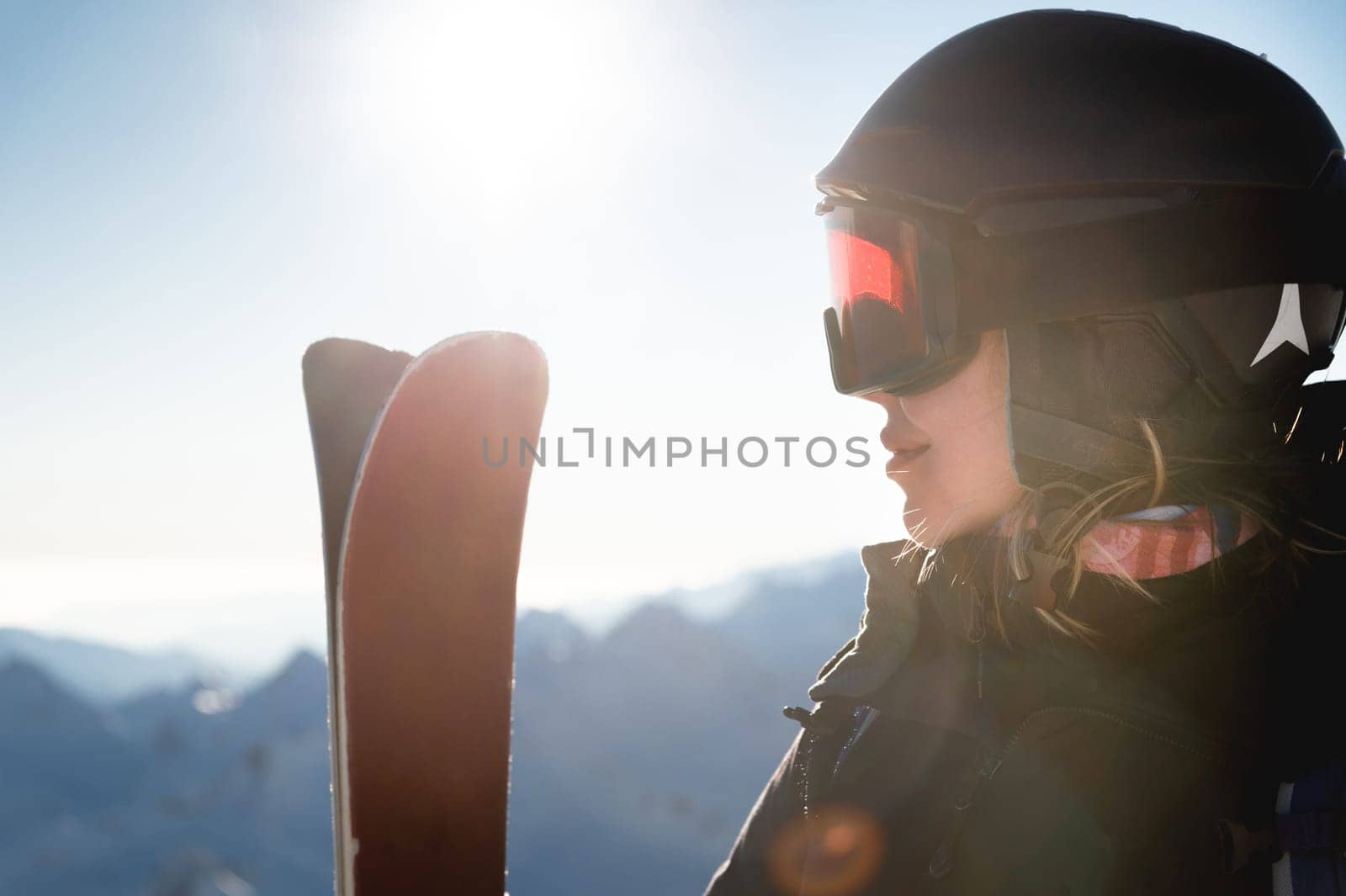 Portrait of sportswoman in helmet and mask with skis in hand, looking away, enjoying sunny frosty day, perfect day for skiing in ski resort.