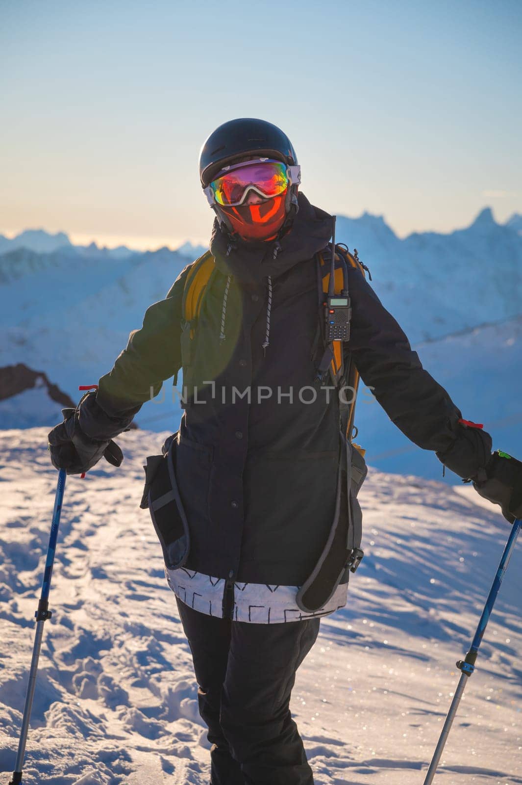 Winter skis. Ski portrait of a woman skier. Portrait of happy skier holding sticks and looking at camera against snowy mountains at sunset in the background.