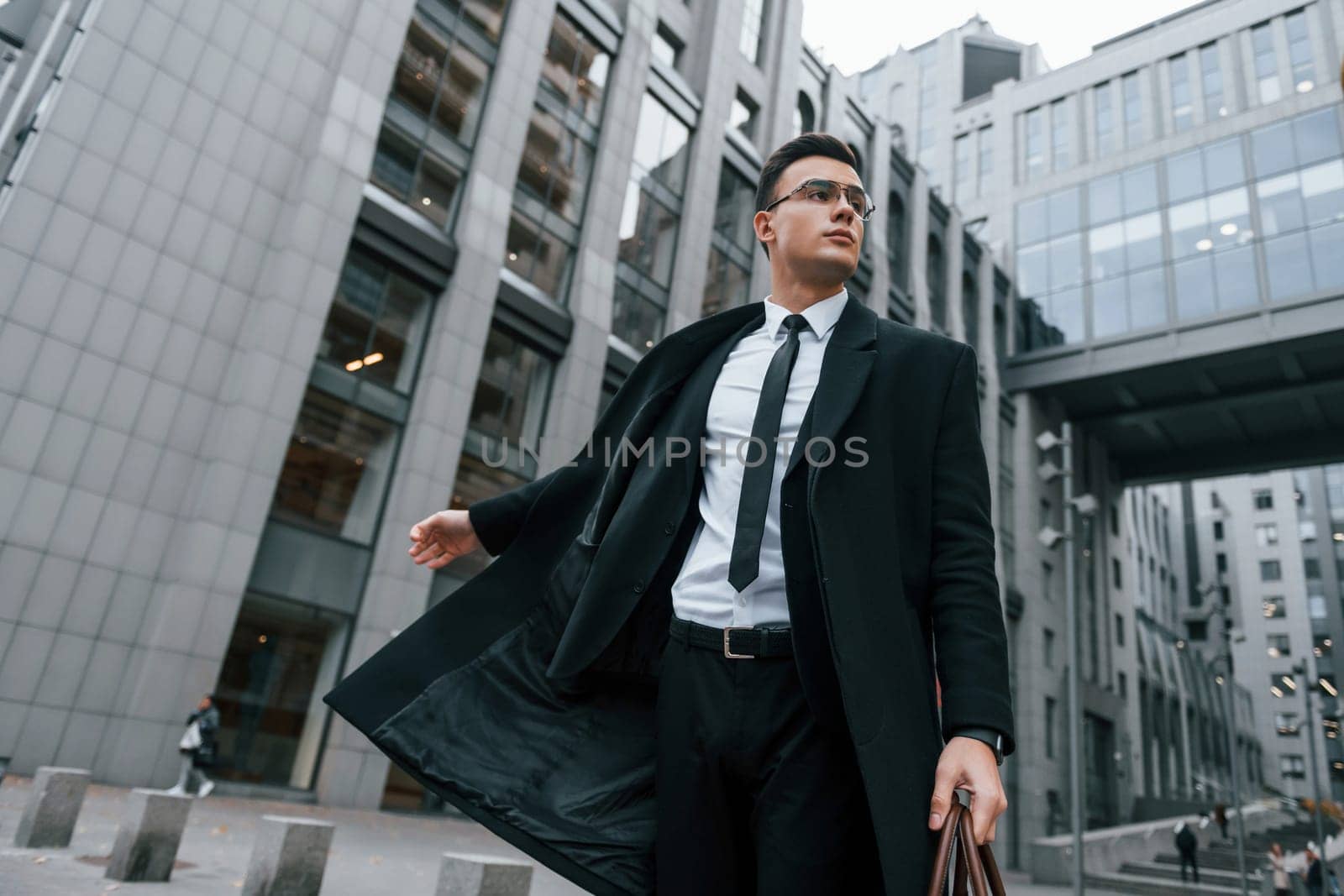 Grey building behind. Businessman in black suit and tie is outdoors in the city by Standret