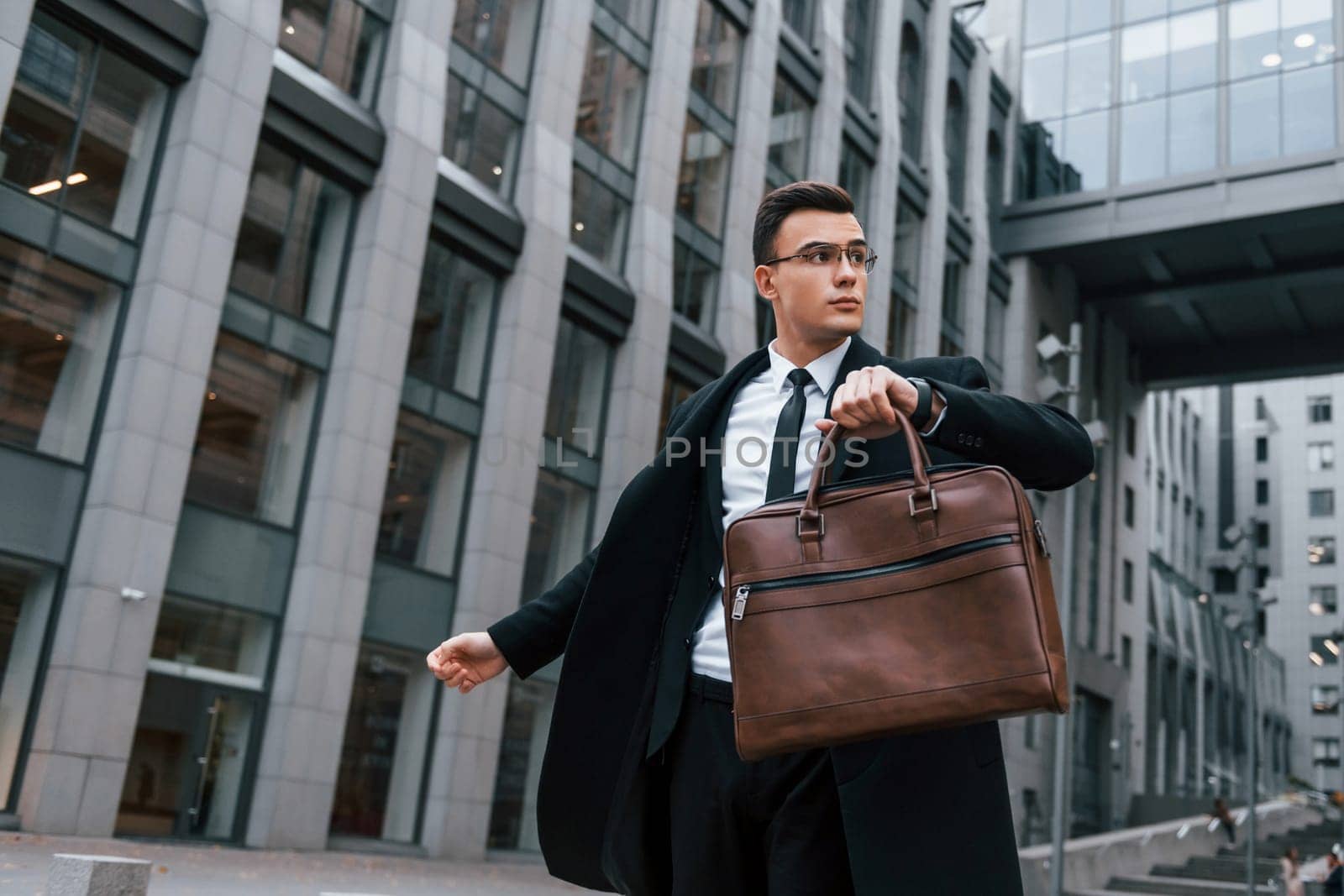 Grey building behind. Businessman in black suit and tie is outdoors in the city.