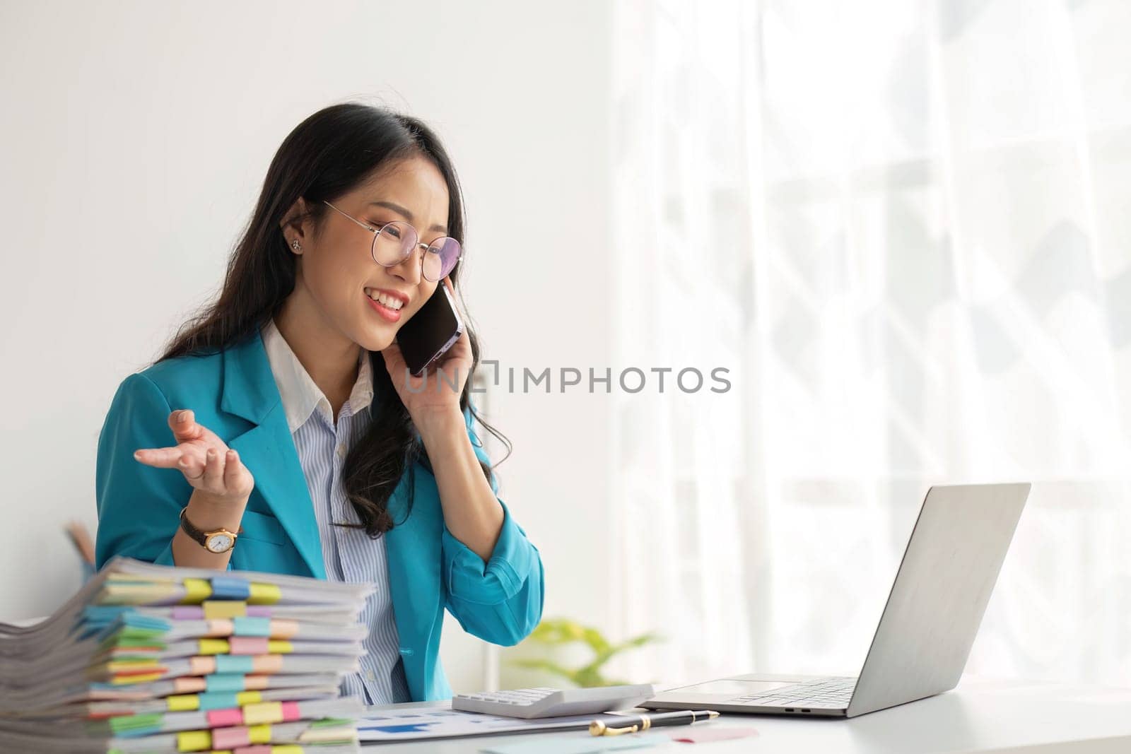 Asian Woman entrepreneur busy with her work in the office. Young Asian woman talking over smartphone or cellphone while working on computer at her desk...