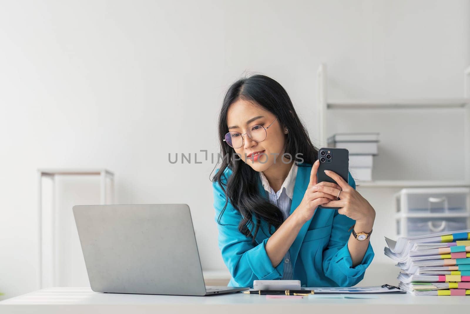 Asian Woman entrepreneur busy with her work in the office. Young Asian woman talking over smartphone or cellphone while working on computer at her desk...