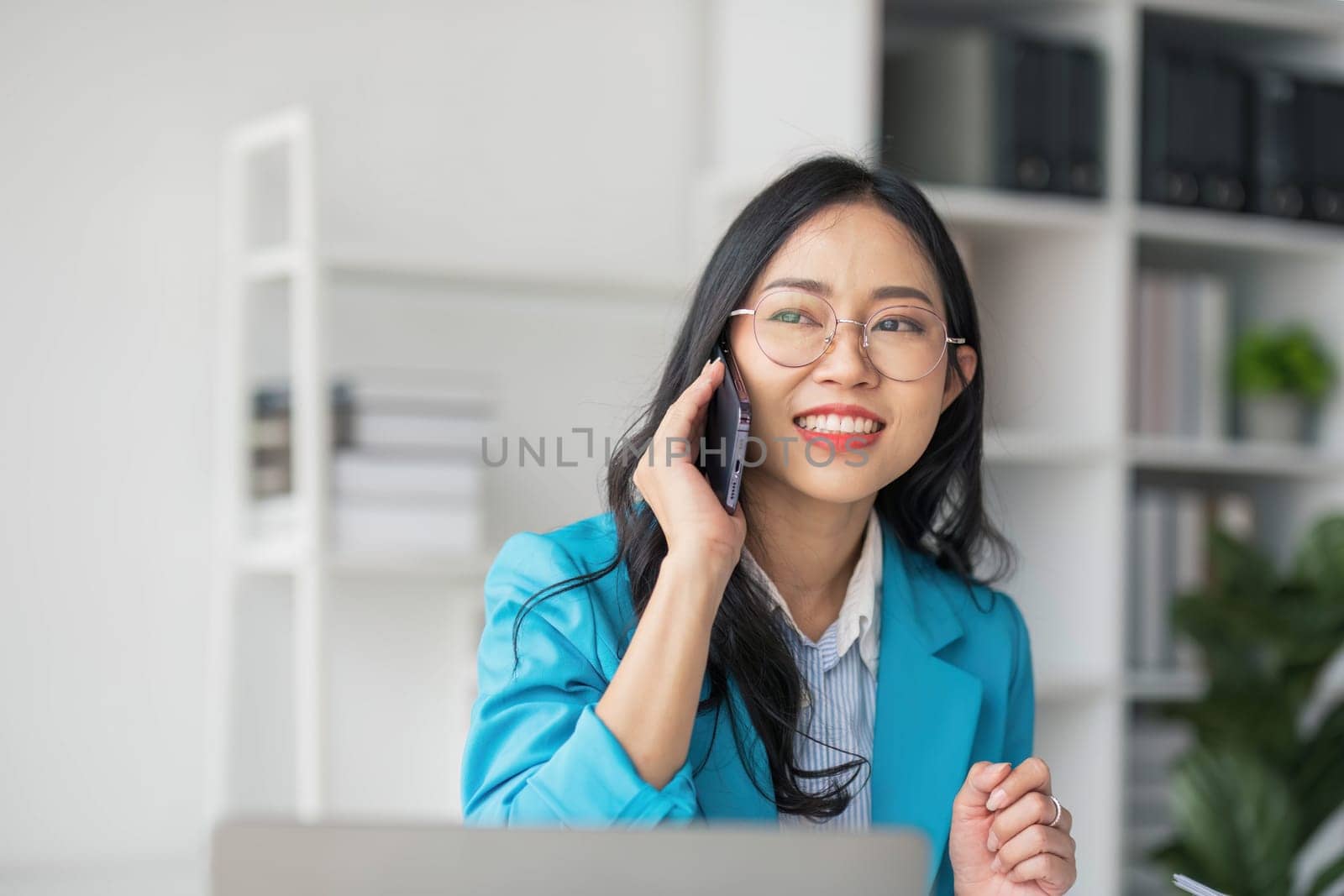 Asian Woman entrepreneur busy with her work in the office. Young Asian woman talking over smartphone or cellphone while working on computer at her desk...