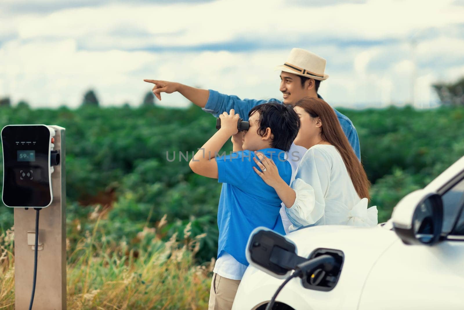 Concept of progressive happy family enjoying their time at wind farm with electric vehicle. Electric vehicle driven by clean renewable energy from wind turbine generator for charging station.