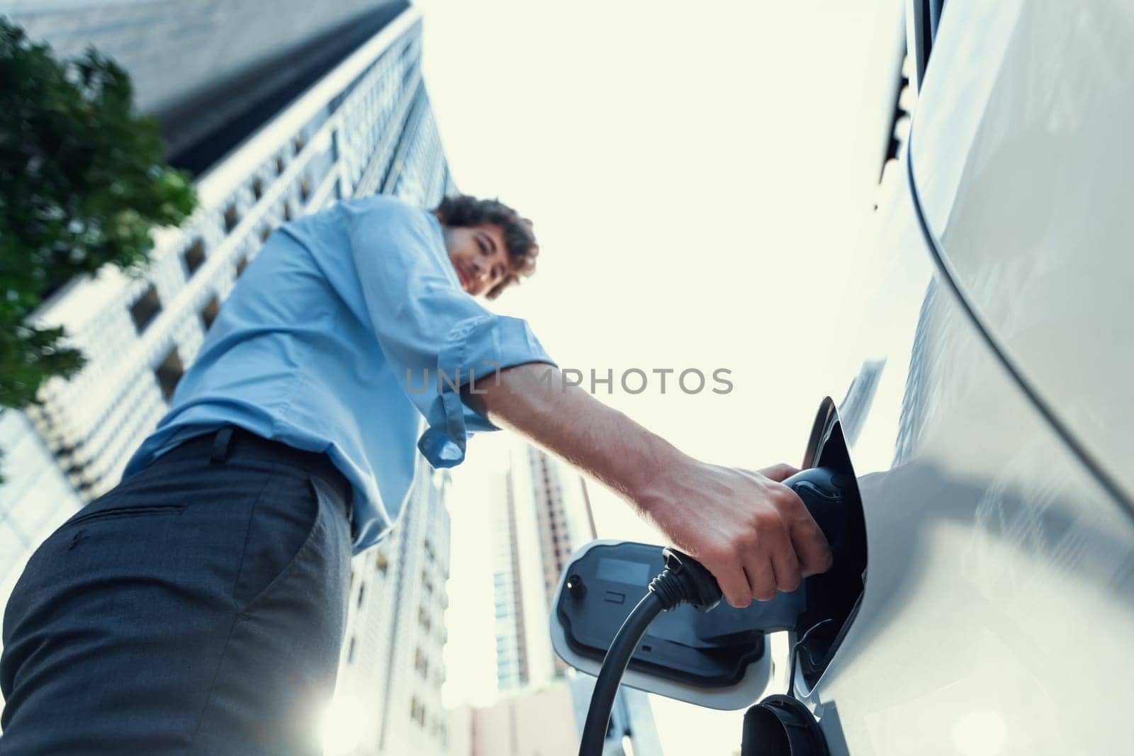 Fisheye view focus on hand insert EV charger plug into electric car with blur background of progressive modern city lifestyle-businessman recharge his EV car with residential building condo background