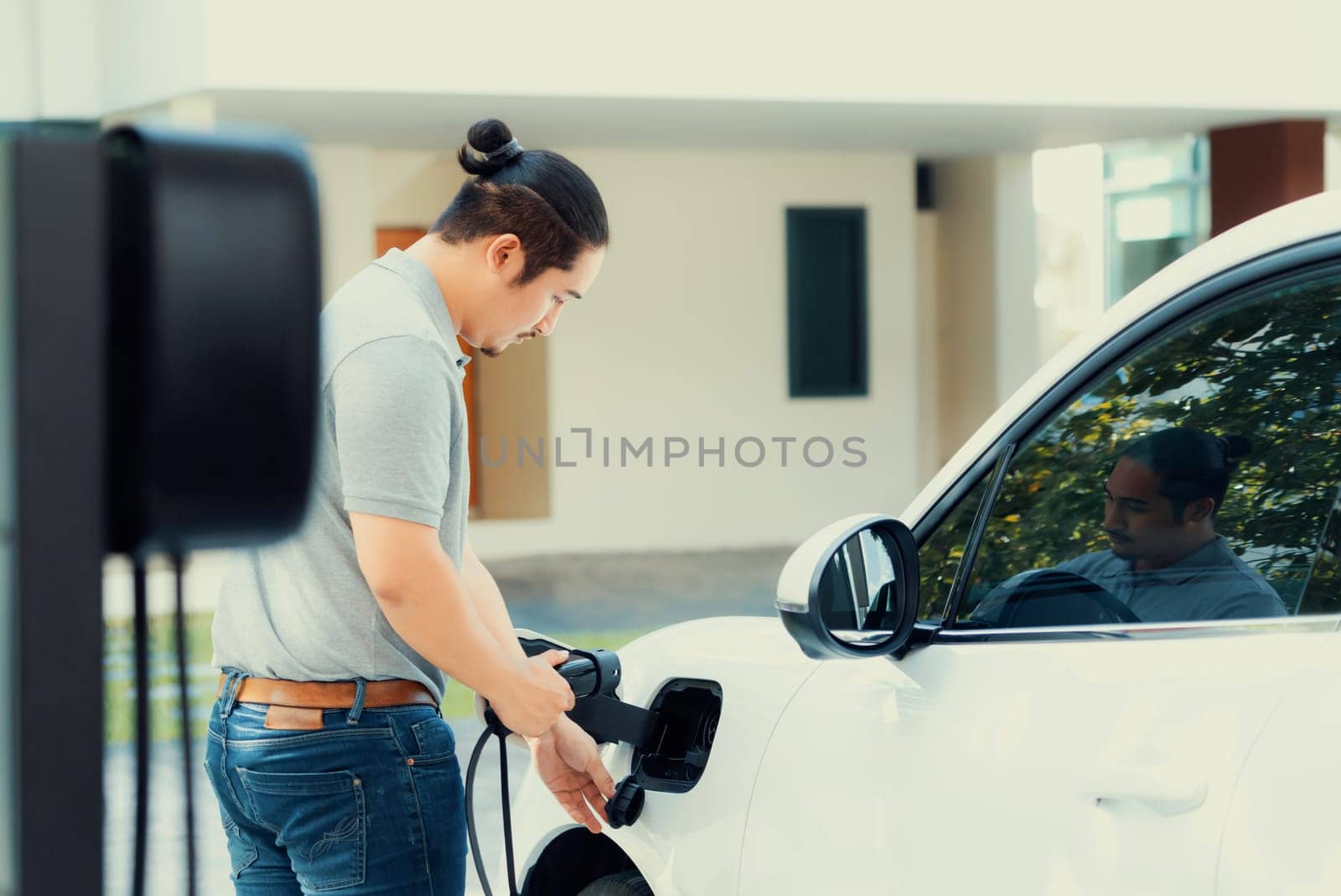 Progressive asian man install cable plug to his electric car with home charging station in the backyard. Concept use of electric vehicles in a progressive lifestyle contributes to clean environment.