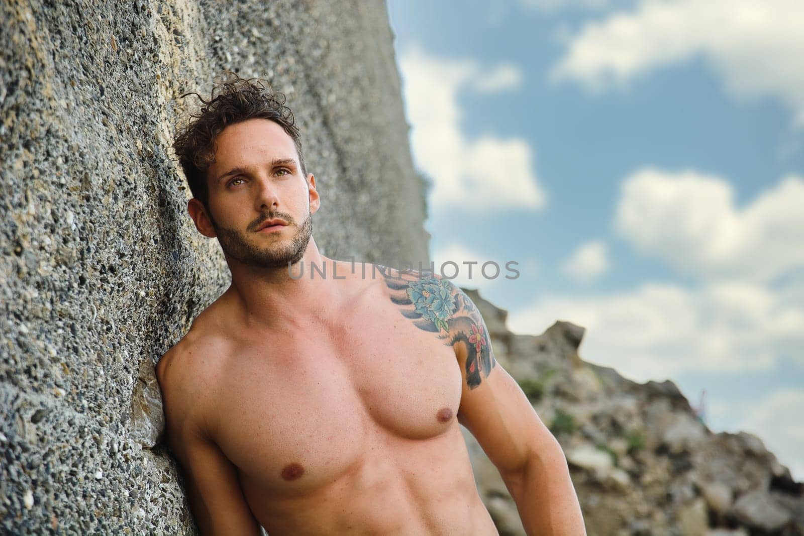 Attractive muscular shirtless athletic man standing on rock next to water by sea or ocean shore, looking at camera in a cloudy summer day