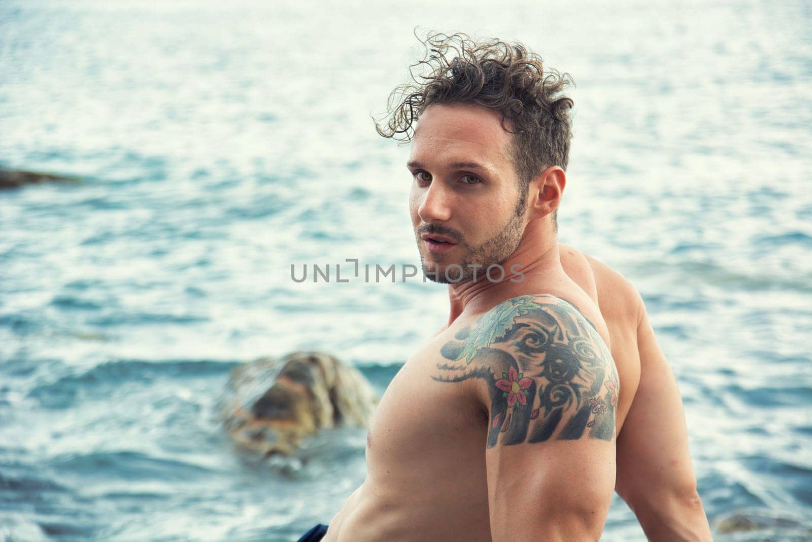 Attractive muscular shirtless athletic man sitting next to water by sea or ocean shore, looking at camera in a cloudy summer day