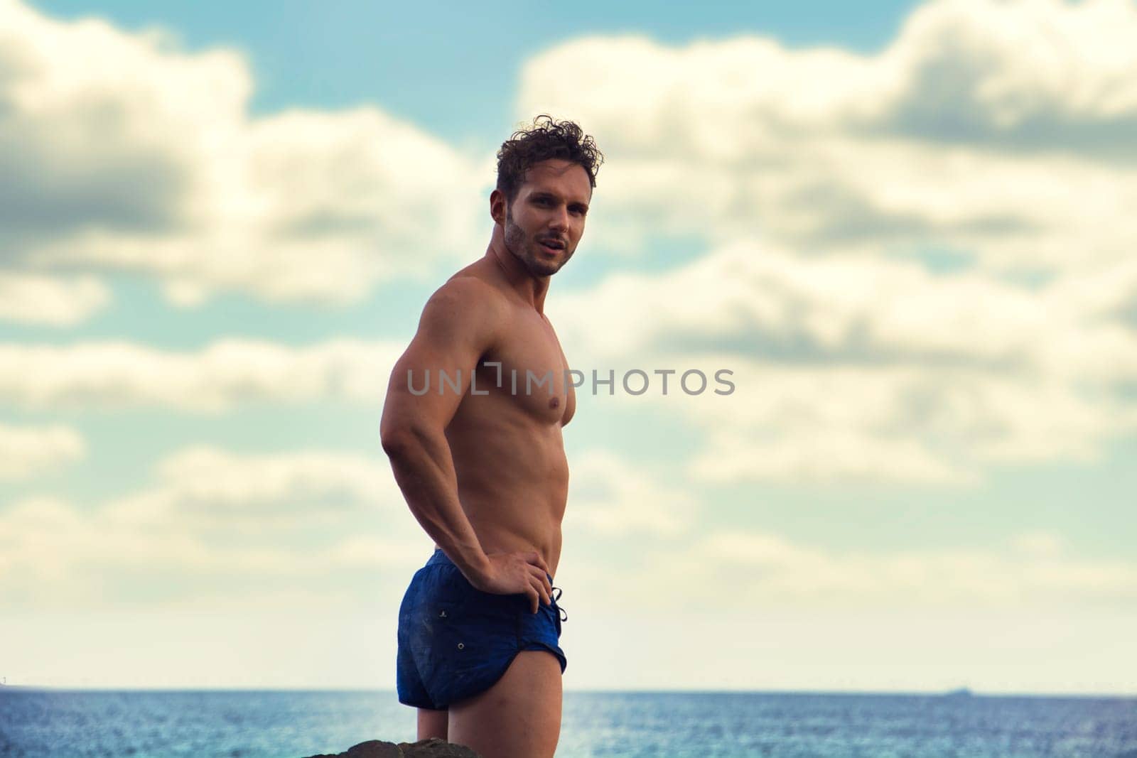 Attractive muscular shirtless athletic man standing next to water by sea or ocean shore, looking at camera in a cloudy summer day