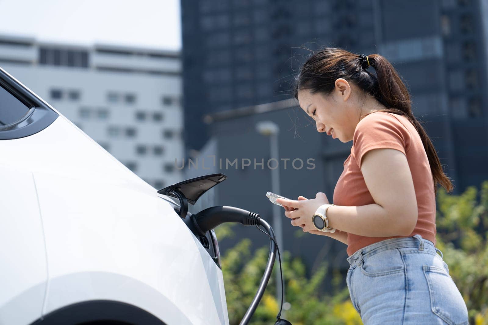 A young woman and her electric car at electric charging station in city center . EV urban lifestyle concept