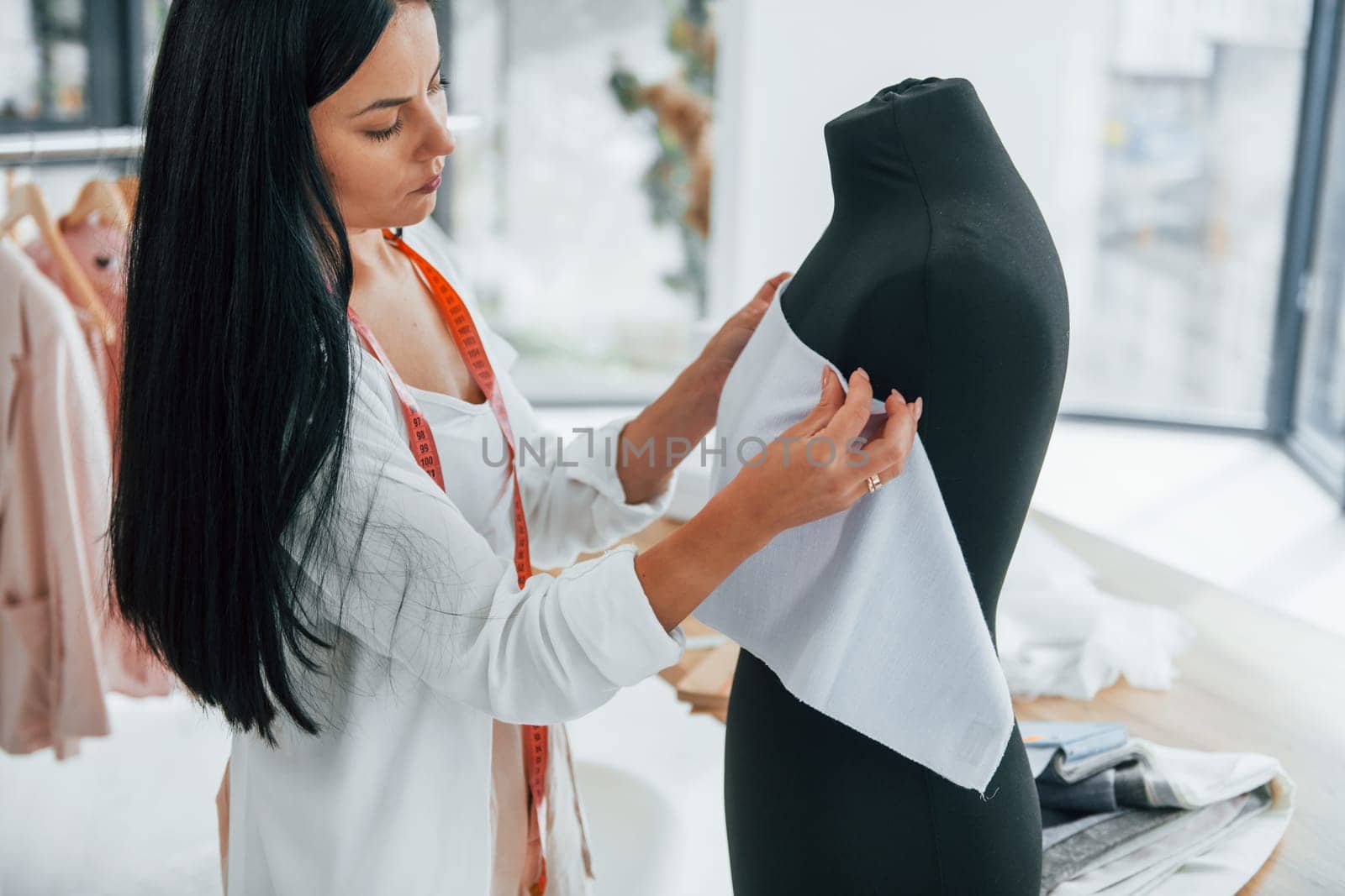 Using measuring tape. Seamstress is in her office that is with different clothes.