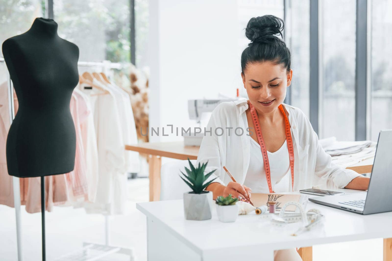 Sitting by the table. Seamstress is in her office that is with different clothes.