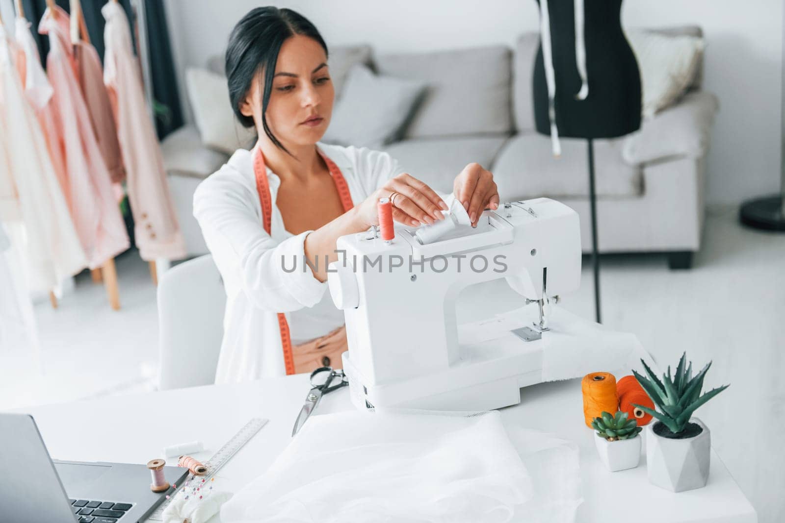 Working by using sewing machine. Seamstress is in her office that is with different clothes.